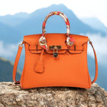Orange leather satchel handbag with scarf-wrapped handle and gold hardware placed on a mountain rock – Aurora Bags outdoor lifestyle shot